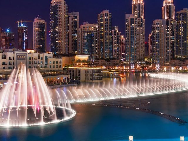 a water fountain with lights and buildings in the background